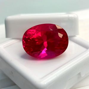 May include: A vibrant, oval-cut red gemstone with a faceted surface that reflects light, displayed resting on a white plastic jewelry box. The stone exhibits a deep, saturated crimson hue and a polished finish, showcasing clear geometric facets.