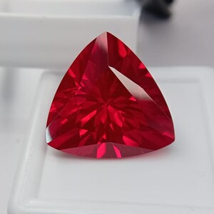 Puede incluir: Una gema de color rojo vibrante tallada en forma triangular con múltiples facetas que crean un patrón de reflexión de luz en forma de estrella. La piedra está colocada sobre una base de exhibición cuadrada blanca, mostrando su color carmesí.