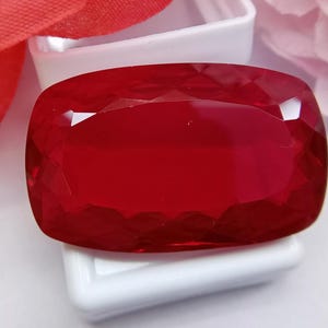 以下が含まれることがあります： ファセットカットが施されたクッションカットの鮮やかな深紅の宝石。石は光を捉える幾何学的なファセットを持つ研磨された反射面を特徴とし、輝きを放っています。小さな白い容器の上に置かれています。