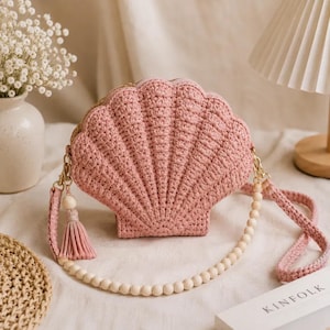 May include: A dusty pink crochet handbag shaped like a seashell. The bag features a textured ribbed pattern, a matching crochet strap, a decorative tassel with a wooden bead, and a long shoulder strap made of smooth, round wooden beads.