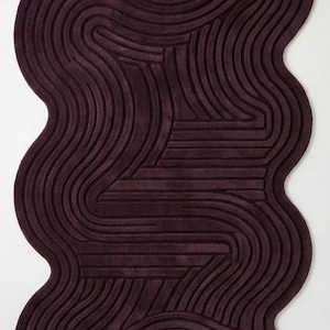 Puede incluir: Una alfombra de color morado oscuro con una forma orgánica, irregular y ondulada. La superficie presenta una serie de líneas en relieve, estilo tallado, que siguen los contornos de la alfombra, creando un patrón lineal texturizado en toda la zona.