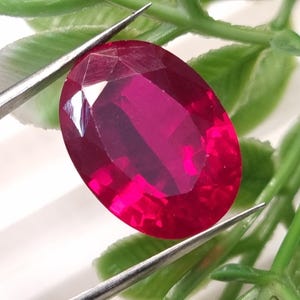 May include: A vibrant, oval-cut, deep red gemstone held by silver-toned metal tweezers. The stone features a faceted surface that reflects light, showcasing a rich, saturated crimson hue throughout its transparent, polished structure.