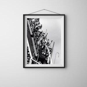 Puede incluir: Una fotografía en blanco y negro en un marco negro que muestra una montaña rusa de madera llamada Cyclone en Coney Island. Varios pasajeros están sentados en los vagones, algunos levantando los brazos mientras recorren la vía empinada.