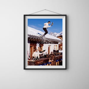 May include: A framed wall art print depicting a skier in a white jacket jumping off a snow-covered roof while holding a tray with glasses. Below, a person in a white suit pours a bottle and another in a fur coat sits at an outdoor bar.