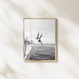 May include: A black and white framed wall art print depicting two individuals jumping from the side of a sailboat into the open ocean. The scene captures the figures mid-air against a bright sky, with the boat's railing visible in the foreground.
