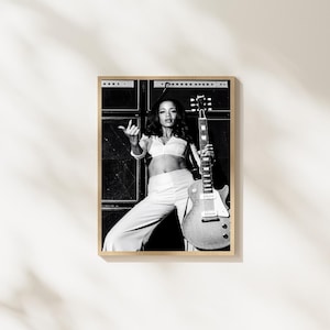 May include: A black and white photograph of a musician wearing a cropped top and trousers, standing in front of stacked guitar amplifiers. The musician holds an electric guitar with the neck pointing upward and gestures with one hand towards the camera.