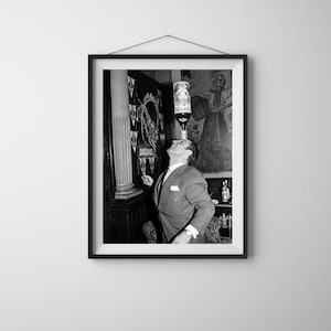May include: A black and white framed photograph showing a person in a suit balancing a Martini bottle on their forehead. The background features a wall with a fluted column and a mural depicting a figure. The bottle label clearly displays the text Martini.