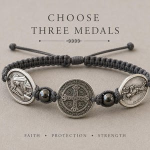 May include: A dark gray cord bracelet featuring three silver-toned metal medals and two dark spherical beads. The central medal displays a cross with text, while the side medals depict religious figures. The text CHOOSE THREE MEDALS is visible.