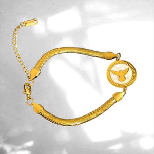 Peut inclure: Un bracelet en chaîne serpent dorée avec un pendentif circulaire incrusté de nacre blanche et une silhouette d'oiseau dorée au centre. Le bracelet est doté d'un fermoir mousqueton et d'une chaînette d'extension avec une petite breloque.