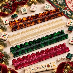 May include: Four long, rectangular mahjong tile racks with scalloped edges, arranged in a stack. The racks feature translucent, marbled patterns in tortoise shell, cream, dark green, and deep red, accompanied by scattered mahjong tiles.