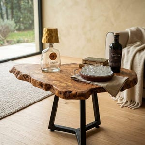 Op de afbeelding: Een houten salontafel met een natuurlijke houtnerf en onregelmatige randen, ondersteund door een zwart metalen driepootonderstel. Op de tafel staan een glazen flessenlamp met een gouden kap, een donkere fles met het label Trentina.