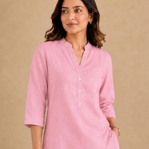 May include: A light pink tunic top featuring a mandarin collar, a V-neckline, and a partial button-down placket with three white buttons. The garment has three-quarter length sleeves and a straight hemline, crafted from a solid-colored fabric.