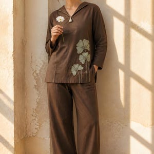 May include: A dark brown long-sleeved tunic top with a V-neckline and a small tassel detail, featuring light green floral embroidery on the lower left side, paired with matching dark brown wide-leg trousers and brown flat sandals.