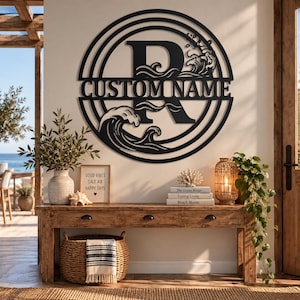 May include: A circular black metal wall sign featuring a large letter R in the center, the text CUSTOM NAME, and stylized ocean wave cutouts. The design is enclosed within concentric circular rings, creating a decorative piece of wall art.
