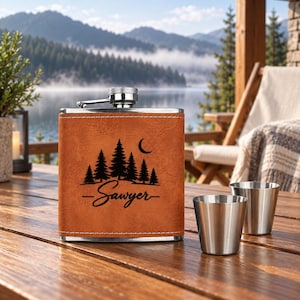 Puede incluir: Una petaca rectangular de acero inoxidable recubierta de piel sintética color canela con un diseño grabado a láser en negro de pinos y una luna creciente sobre el nombre Sawyer en cursiva. Dos vasos pequeños de acero inoxidable al lado.