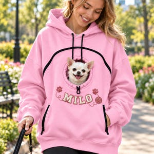 Könnte beinhalten: Ein hellrosa Kapuzenpullover mit einer mittigen, runden Tasche mit Kordelzug, die zum Tragen eines kleinen Hundes dient. Der Taschenbereich ist mit dem Namen MILO, Pfotenabdrücken und einer Wollknäuel-Grafik sowie schwarzen Akzenten bestickt.