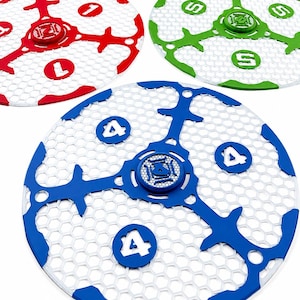 Op de afbeelding: Drie ronde spelschijven met een witte honingraatgaasbasis. Eén schijf is rood met het cijfer 1, één is groen met de letter S en één is blauw met het cijfer 4. Elke schijf heeft een centraal embleem en drie uitstralende armen.