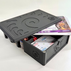 Könnte beinhalten: Eine schwarze, rechteckige Aufbewahrungsbox für Spielkarten mit einer Schublade und einem Deckel mit Spiralmuster. Die Box enthält Spielkartenstapel, darunter eine mit der Aufschrift Ravager, sowie kleine rote Würfel und ein Wabenmuster.