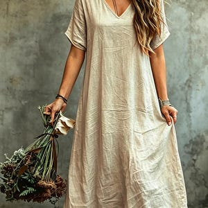 K&ouml;nnte beinhalten: Ein beiges, kurz&auml;rmeliges Maxikleid mit V-Ausschnitt aus strukturiertem Stoff. Das Kleid hat eine lockere, flie&szlig;ende Silhouette, die bis zu den Kn&ouml;cheln reicht. Das Kleidungsst&uuml;ck besticht durch ein schlichtes Design mit sichtbaren Falten im Stoff.
