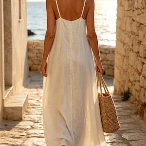 Pu&ograve; includere: Un abito lungo senza maniche color bianco sporco con spalline sottili e una silhouette semplice e fluida. L'abito presenta uno scollo dritto sulla schiena e una cucitura verticale al centro, abbinato a una borsa tote in paglia intrecciata.