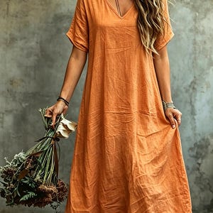 K&ouml;nnte beinhalten: Ein langes, locker sitzendes orangefarbenes Kleid mit V-Ausschnitt und kurzen &Auml;rmeln. Das Kleidungsst&uuml;ck hat eine texturierte, leicht knittrige Stoffoberfl&auml;che und reicht bis zur Waden- oder Kn&ouml;chell&auml;nge, was eine entspannte Silhouette ergibt.