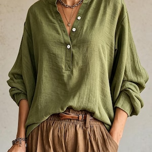 May include: An olive green long-sleeve shirt with a mandarin collar, a partial button-down placket with three white buttons, and loose-fitting sleeves. The shirt is paired with brown trousers featuring a brown leather belt at the waist.