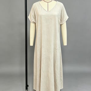 K&ouml;nnte beinhalten: Ein beiges Maxikleid mit V-Ausschnitt und kurzen &Auml;rmeln. Das Kleid hat eine lockere, flie&szlig;ende Silhouette, die bis zu den Kn&ouml;cheln reicht, und ist aus einem einfarbigen Stoff mit dezenter Textur gefertigt, pr&auml;sentiert auf einem Schneiderb&uuml;stenst&auml;nder.