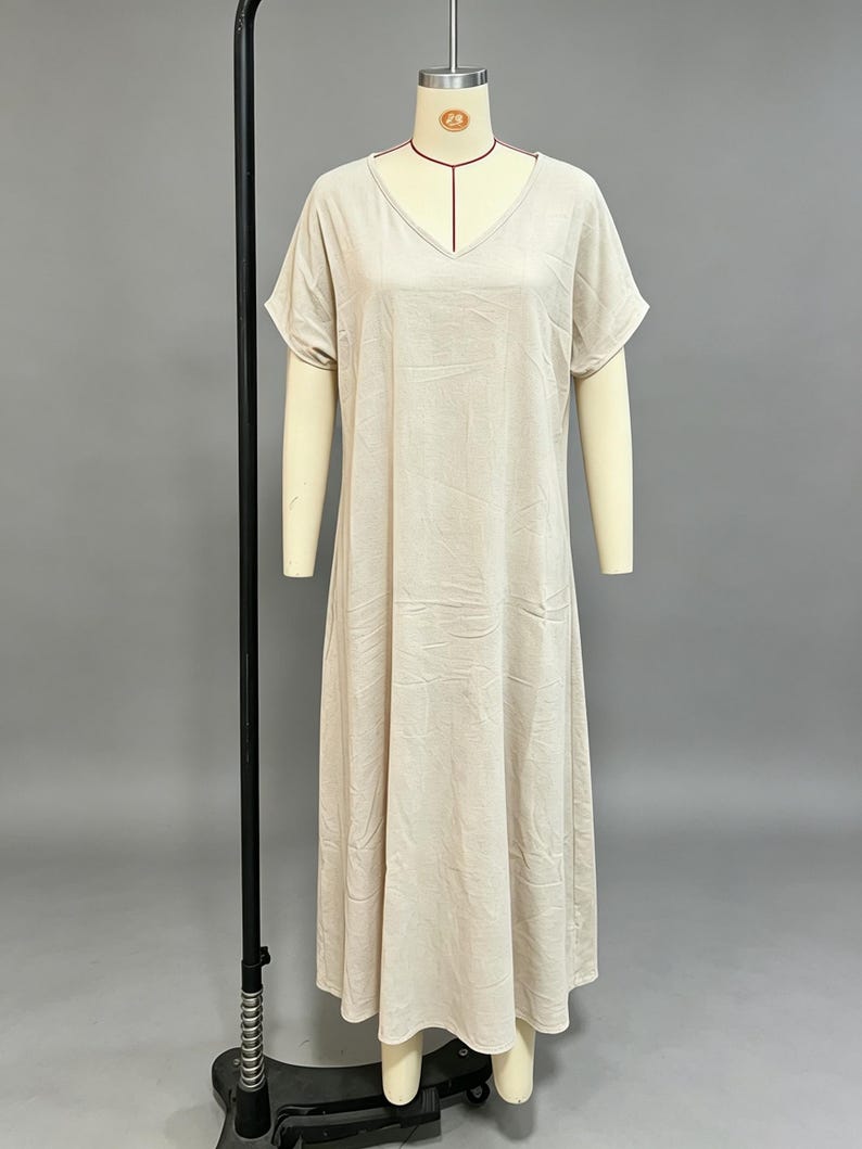 K&ouml;nnte beinhalten: Ein beiges Maxikleid mit V-Ausschnitt und kurzen &Auml;rmeln. Das Kleid hat eine lockere, flie&szlig;ende Silhouette, die bis zu den Kn&ouml;cheln reicht, und ist aus einem einfarbigen Stoff mit dezenter Textur gefertigt, pr&auml;sentiert auf einem Schneiderb&uuml;stenst&auml;nder.