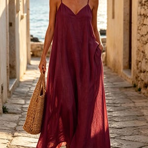 Pu&ograve; includere: Un abito lungo color bordeaux intenso con spalline sottili, scollo a V e una silhouette ampia e fluida con tasche laterali. Il capo &egrave; abbinato a una borsa tote in paglia intrecciata color cuoio e a sandali aperti.