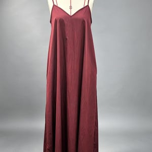 Pu&ograve; includere: Un abito lungo color bordeaux intenso con scollo a V e doppi spallacci sottili. Il capo presenta una silhouette ampia e fluida con una finitura in tessuto liscio e tinta unita, esposto su un manichino su uno sfondo neutro.