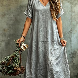K&ouml;nnte beinhalten: Ein hellgraues Midikleid mit V-Ausschnitt und kurzen &Auml;rmeln. Das Kleidungsst&uuml;ck hat eine lockere, flie&szlig;ende Silhouette und eine strukturierte Stoffoberfl&auml;che. Das Kleid wird mit einem Strau&szlig; getrockneter Blumen und braunen Lederstiefeln kombiniert.