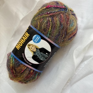 May include: A ball of fuzzy, multi-colored mohair yarn featuring streaks of green, brown, and pink with metallic silver accents. A black paper label wraps around the center, displaying the text MOHAIR, MACHINE WASHABLE, and FREE PATTERN INSIDE.