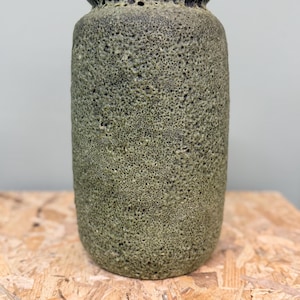May include: A cylindrical ceramic vase with a textured, moss-green exterior featuring a porous, sponge-like surface finish. The vase has a dark, smooth, rounded rim at the top and a straight body that tapers slightly toward the base, creating a sturdy form.