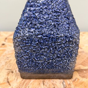 Puede incluir: Un jarrón de cerámica de forma cuadrada con una superficie muy texturizada y rugosa en tonos azul oscuro y negro. La base del jarrón es lisa y sin esmaltar, mostrando un tono de arcilla marrón natural, mientras que el cuerpo superior tiene un esmalte grueso.