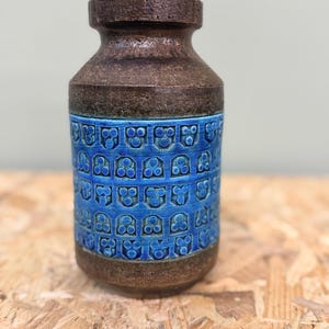 May include: A cylindrical ceramic vase featuring a dark brown, textured top and base with a wide, vibrant blue central band. The blue section is decorated with a repeating embossed pattern of small, stylized geometric shapes arranged in horizontal rows.