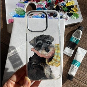 May include: A clear phone case featuring a hand-painted illustration of a black and grey Schnauzer dog. The dog is depicted with a tan and yellow plaid scarf around its neck and a black collar with a gold buckle. The artwork is detailed and textured.