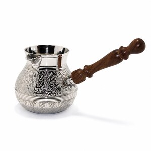 May include: A silver-colored metal coffee pot with a wide, rounded body and a long, dark brown wooden handle. The exterior of the pot features intricate embossed floral and leaf patterns, with a smooth, polished rim and a small pouring spout.