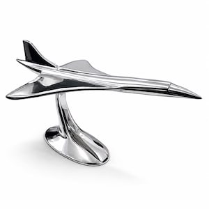 Könnte beinhalten: Ein poliertes, silberfarbenes Metallmodell eines Überschall-Düsenflugzeugs. Die Skulptur verfügt über einen schlanken, aerodynamischen Rumpf mit Deltaflügeln und einem Seitenleitwerk, montiert auf einem geschwungenen, ovalen Sockel.