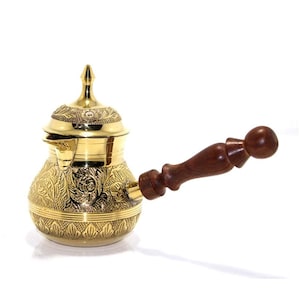 May include: A polished brass coffee pot featuring an intricate floral and leaf engraving across its rounded body and lid. The vessel includes a pointed spout and a long, dark brown wooden handle with a turned, decorative shape for a firm grip.