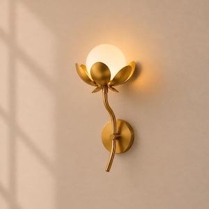 Könnte beinhalten: Eine Wandleuchte mit einem goldfarbenen Metallstiel und einer blütenförmigen Basis mit geschwungenen Blättern, die eine kugelförmige, mattweiße Glühbirne umschließen. Die Leuchte ist an einer runden, goldfarbenen Metallplatte befestigt.