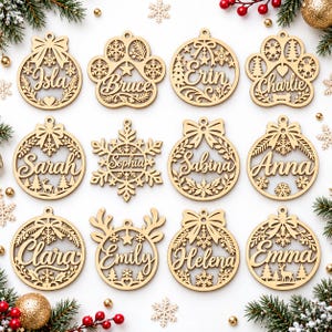 Puede incluir: Una colección de doce adornos navideños de madera cortados con láser que presentan intrincados diseños festivos como copos de nieve, lazos y huellas de patas. Cada adorno está personalizado con un nombre: Isla, Bruce, Erin, Charlie, Sarah, Sophia, Sabina, Anna, Clara, Emily, Helena y Emma.