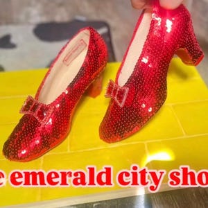 May include: A pair of red sequined pumps with a small block heel and a decorative bow on the toe. The shoes are placed on a yellow rectangular base with a brick pattern. The text "The emerald city shop" is printed in red at the bottom.