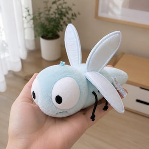 May include: A light blue plush toy shaped like a dragonfly with large white eyes, black pupils, and thin black legs. The toy features two pairs of upright, pale blue wings and a small tuft of blue fuzz on its head, crafted from soft fabric.