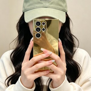 May include: A rectangular smartphone case with a highly reflective gold mirror finish. The case features a cutout section for a multi-lens camera array on the upper left side, showing four circular lenses and a small flash component in a vertical layout.