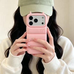 May include: A light pink, puffer-style phone case with a soft, quilted texture and two small, pointed ears at the top. The case features a white line-art graphic of two stick figures and the word "Fabulous" embossed near the bottom edge.