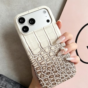 May include: A smartphone case featuring a textured, cream-colored faux crocodile skin pattern that transitions into a darker brown, scale-like texture at the bottom. A small, decorative white bow with a dangling crystal accent is attached to the surface.