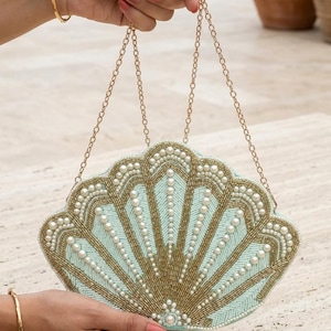 May include: A shell-shaped clutch bag featuring light blue and gold beadwork arranged in a radial pattern, accented with white faux pearls along the edges and centre. The bag includes a thin gold-tone metal chain strap for carrying.