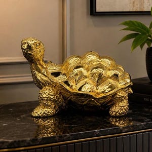 May include: A decorative turtle figurine with a highly reflective, polished gold finish. The sculpture features a textured head and legs with a smooth, segmented shell design. The turtle is positioned in a forward-facing stance, showcasing a metallic lustre that catches the light, creating a bright and prominent appearance.