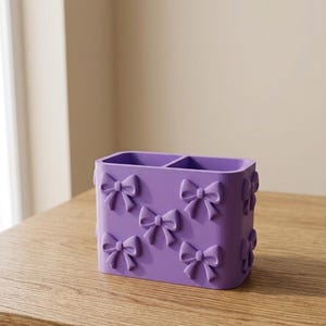 Può includere: Un organizer da scrivania rettangolare color lavanda con due scomparti, caratterizzato da un motivo ripetitivo di fiocchi tridimensionali in rilievo sulla superficie esterna. Il contenitore è realizzato in materiale solido, liscio e arrotondato.