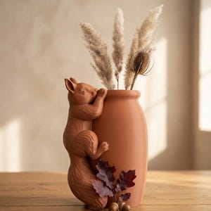 Peut inclure: Un vase en céramique de couleur terre cuite orné d'un écureuil sculpté grimpant sur le côté. Le vase contient des herbes de la pampa séchées et une cardère. Deux petits glands et un groupe de feuilles de chêne rouge foncé sont à la base.
